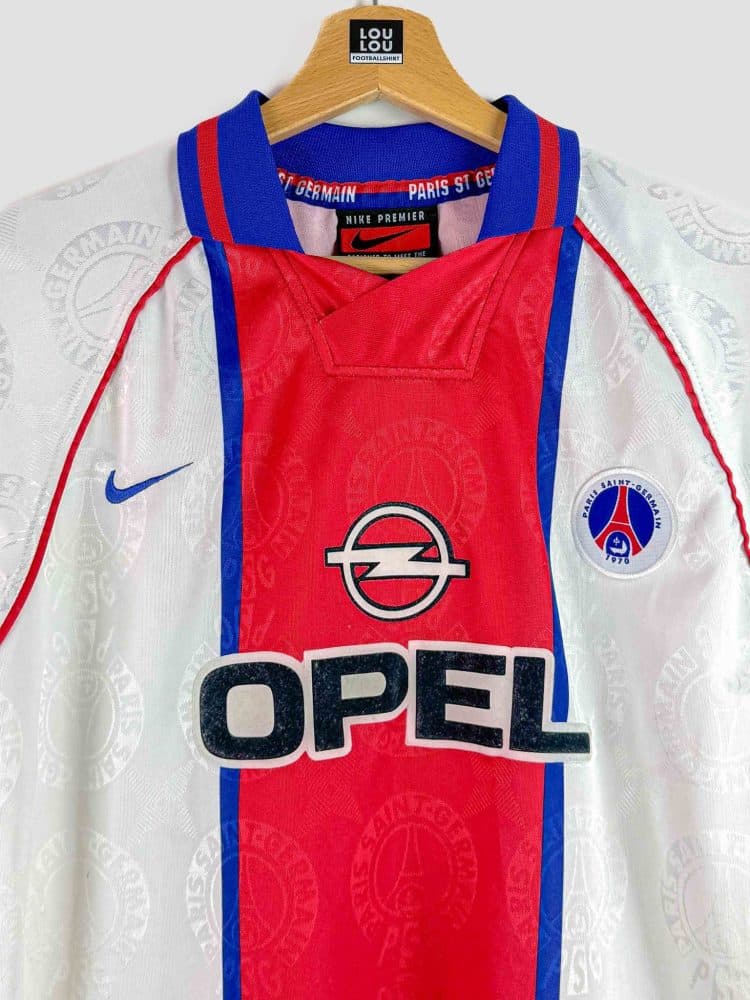 Maillot de collection du Paris SG 96-97 en photographie, design blanc à large bande rouge sur le devant, avec le sponsor Opel affiché au centre et le logo Nike brodé – un classique des années 90.